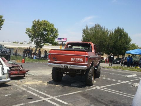 1978 Ford Bronco - A lifted orange pickup truck is parked in a lot next to a damaged vehicle and other cars.