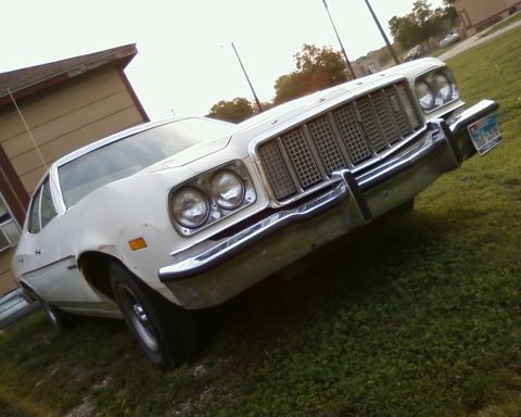 1976 Ford Torino - A white 1970s Ford sedan is viewed from a low angle, highlighting its prominent grille and chrome bumper.