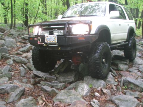 1996 Ford Bronco - This white SUV is shown off-roading on a challenging rocky path surrounded by trees.