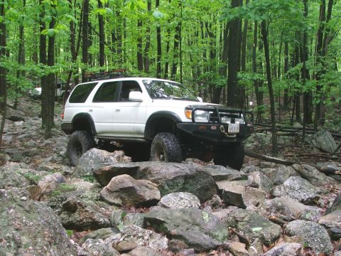 1996 Ford Bronco - A white SUV with aggressive off-road tires is shown navigating a challenging rocky terrain in a dense forest.