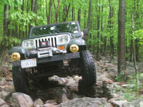 1996 Ford Bronco - A green Jeep Wrangler tackles a challenging rocky path in a wooded area, equipped with a winch and distinctive smiley face decals.