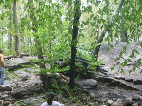 1996 Ford Bronco - Off-road vehicle on a challenging rocky trail surrounded by trees. Two people observe the action.