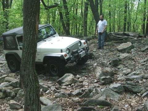 1996 Ford Bronco - A white Jeep Wrangler navigates a rocky trail in a wooded area.