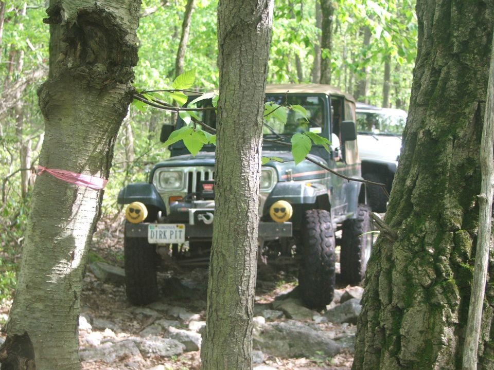 Green Jeep Wrangler with smiley face lights on front, another vehicle behind it, in a wooded area