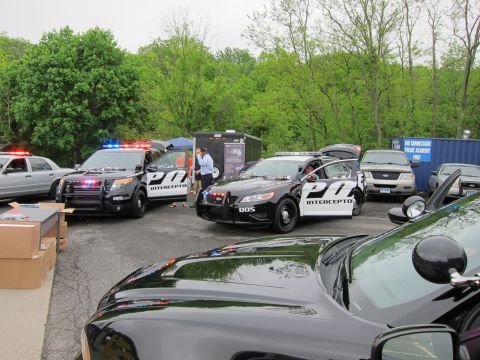 1991 Ford LTD Crown Victoria - group of black-and-white Police Interceptor cruisers with flashing light bars displayed in a paved lot ringed by trees.