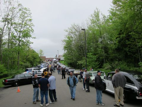 1991 Ford LTD Crown Victoria - crowded outdoor car show under gray skies, with people and rows of parked cars lining a tree-bordered paved drive.