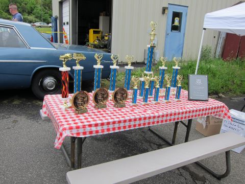 1991 Ford LTD Crown Victoria - lineup of gold trophies and plaques on a red-checkered table outdoors, with a blue vintage sedan and garage behind.