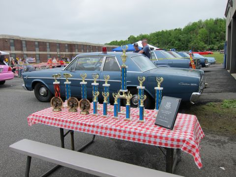 1991 Ford LTD Crown Victoria - Gold trophies arranged on a red-checkered tablecloth, with a blue vintage Ford sedan and more show cars in the background.