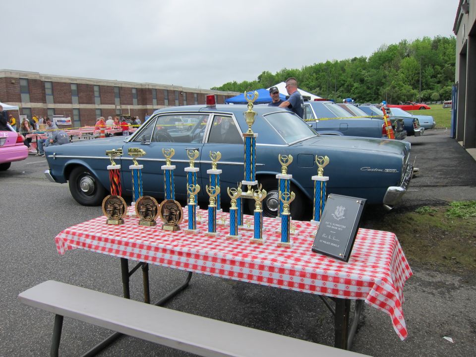 Car-show trophies on a checkered table with a blue vintage Ford sedan behind