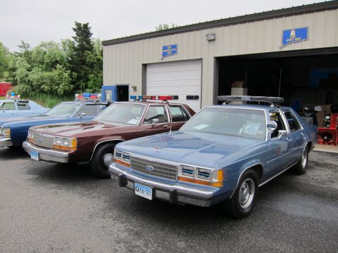 1991 Ford LTD Crown Victoria - row of vintage police sedans, including a blue Ford in front, parked outside a metal garage building.