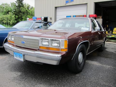 1991 Ford LTD Crown Victoria - maroon Ford LTD Crown Victoria State Police cruiser with a roof light bar parked near a garage at an event.