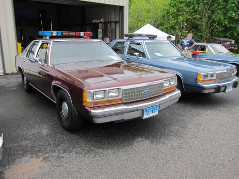 1991 Ford LTD Crown Victoria - maroon Ford State Police sedan parked beside a blue cruiser, with spectators gathered near a tent in the background.