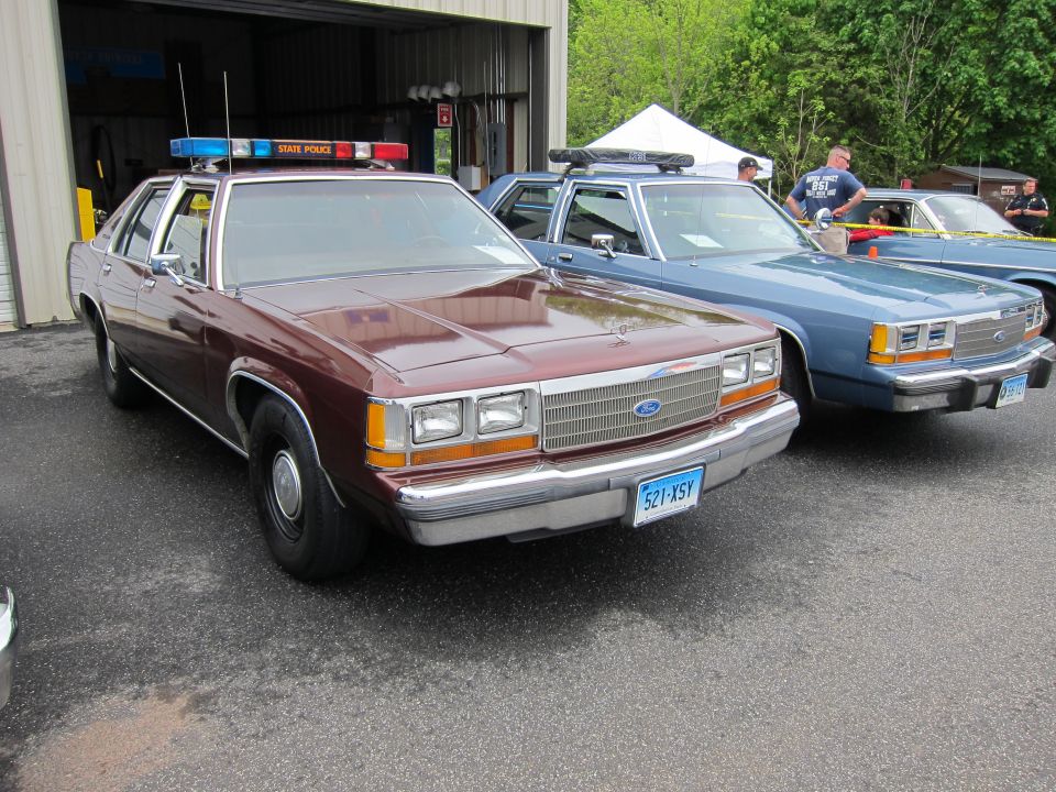 Maroon Ford State Police sedan beside blue cruiser with spectators in background