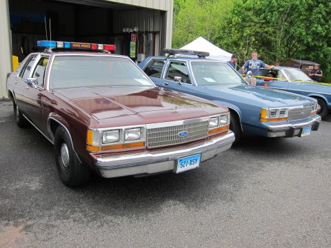 1991 Ford LTD Crown Victoria - maroon Ford State Police sedan and a blue cruiser parked side by side near an open garage bay.