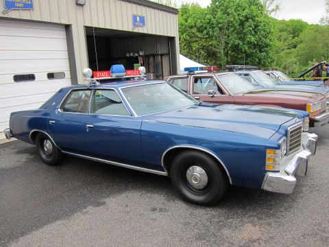 1991 Ford LTD Crown Victoria - blue Ford LTD State Police sedan with red and blue roof beacons parked among other vintage cruisers near a garage.