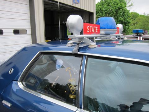 1991 Ford LTD Crown Victoria - Close-up of a blue police car roof showing a red STATE-marked light bar, beacons, and a door-window decal.