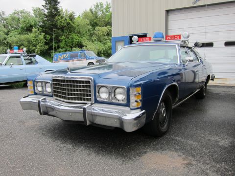 1991 Ford LTD Crown Victoria - blue Ford LTD State Police sedan with red roof beacons parked outside a garage among other vintage cruisers.