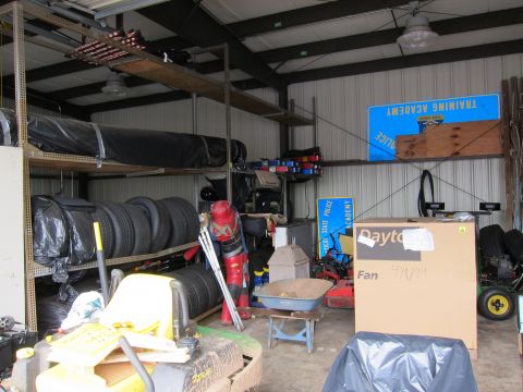 1991 Ford LTD Crown Victoria - Interior of a metal storage shed packed with stacked tires, covered gear, a wheelbarrow, and a boxed fan.