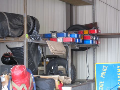 1991 Ford LTD Crown Victoria - Several red and blue police light bars stored on steel shelving inside a metal building beside a State Police academy sign.