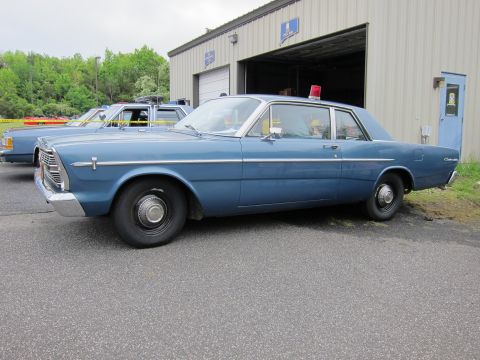 1991 Ford LTD Crown Victoria - Side view of a blue Ford Custom two-door sedan with a roof beacon parked outside a metal garage building.