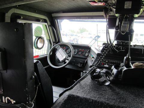 1991 Ford LTD Crown Victoria - Driver's compartment of an armored vehicle showing the steering wheel, instrument gauges, dashboard switches, and a coiled radio handset.