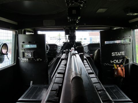 1991 Ford LTD Crown Victoria - Centered rear view inside an armored police truck showing a long mounted firearm on a center rack between padded benches and STATE POLICE s…