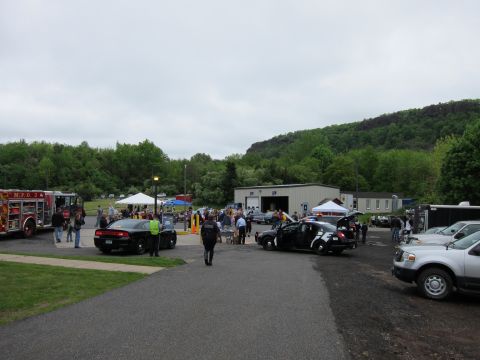 1991 Ford LTD Crown Victoria - Overview of an outdoor public-safety event with a crowd, a fire engine, and police patrol vehicles parked on a paved lot below a wooded rid…
