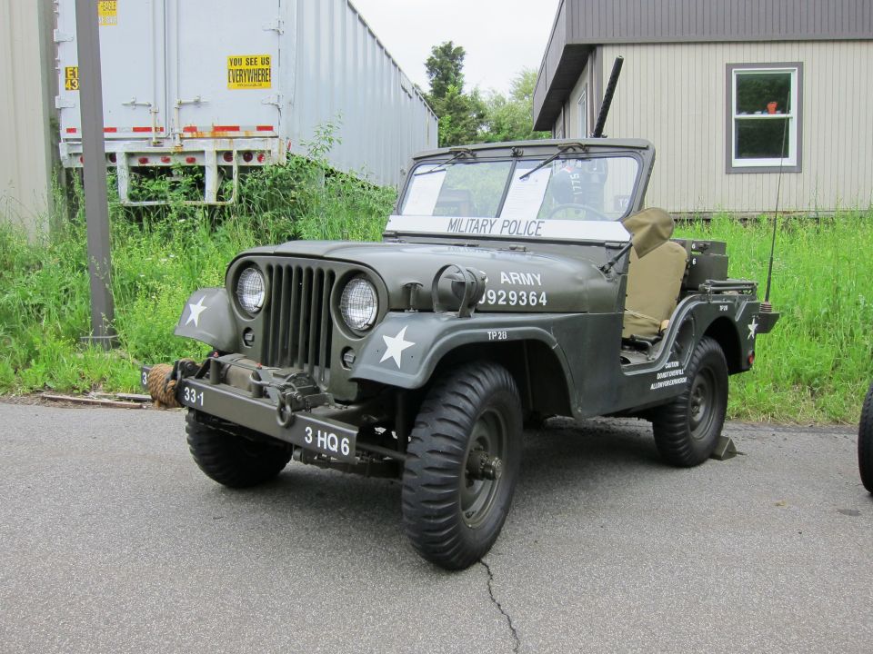 Olive-drab military jeep marked US ARMY MILITARY POLICE parked on pavement by a trailer