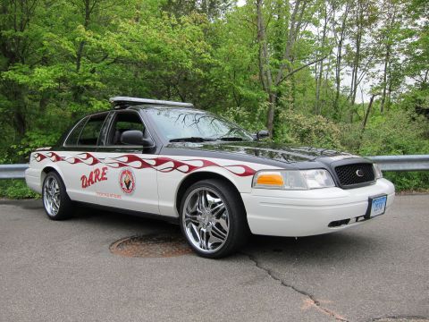 1991 Ford LTD Crown Victoria - white Ford Crown Victoria with dark-red flame graphics, DARE lettering, roof lightbar, and large chrome wheels.