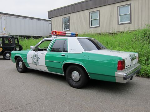 1991 Ford LTD Crown Victoria - Rear three-quarter view of a green-and-white sheriff sedan with roof lightbar and star emblem, parked beside a metal building.