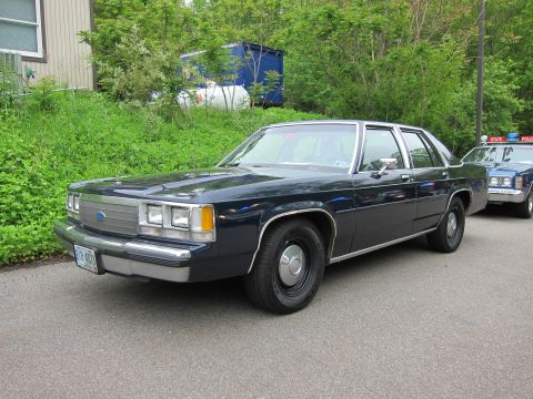 1991 Ford LTD Crown Victoria - Front three-quarter view of a dark blue Ford sedan with chrome trim, parked beside another police vehicle.