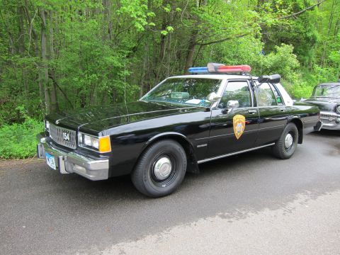 1991 Ford LTD Crown Victoria - black Chevrolet Caprice police sedan numbered 154 with a gold door shield and red-and-blue roof lightbar.