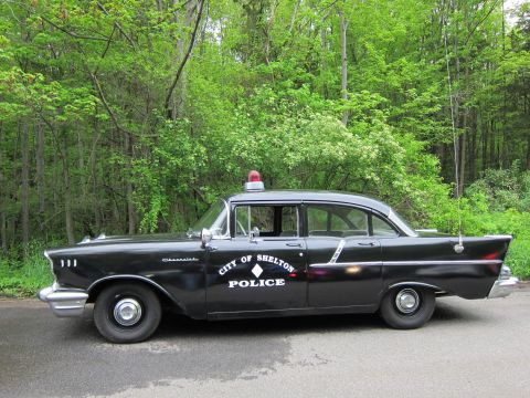 1991 Ford LTD Crown Victoria - Side profile of a black 1957 Chevrolet police sedan lettered CITY OF SHELTON POLICE with chrome trim and red roof beacon.