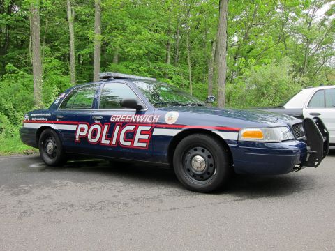 1991 Ford LTD Crown Victoria - Side profile of a dark blue Greenwich Police Ford Crown Victoria with light bar and push bar, parked beside trees.