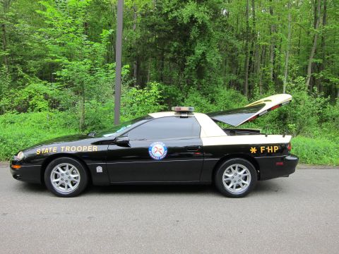 1991 Ford LTD Crown Victoria - black-and-white Chevrolet Camaro marked State Trooper and FHP with light bar, parked on a tree-lined road.