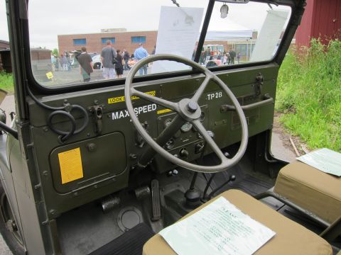 1991 Ford LTD Crown Victoria - Olive-drab military jeep interior showing the steering wheel, dash stencils MAX SPEED and TP 28, and canvas seats, with crowd visible outsi…