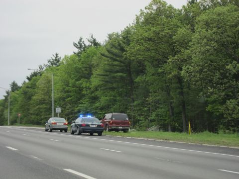 1991 Ford LTD Crown Victoria - police car with activated blue lights stopped behind a sedan and maroon SUV along a tree-lined highway shoulder.