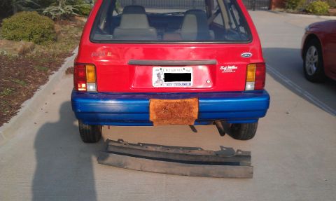 1991 Ford Festiva - The rear of a red and blue 1991 Ford Festiva is shown, with a detached bumper lying in front of it.