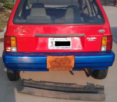 1991 Ford Festiva - Rear view of a 1991 Ford Festiva with a detached bumper trim piece on the ground.