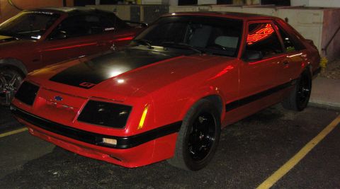 2007 Ford Mustang - This red 1980s Ford Mustang GT features a black hood stripe and black wheels, parked next to another red Mustang.
