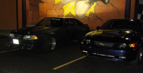 2007 Ford Mustang - Two Ford Mustangs are parked side-by-side in front of a colorful mural at night.