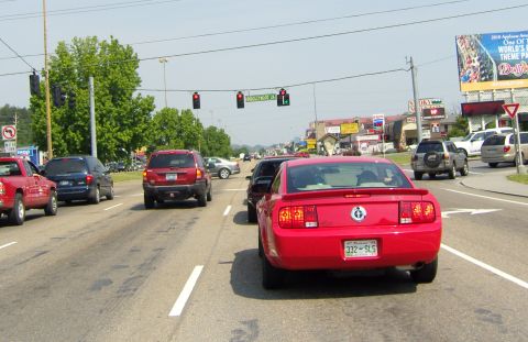 2007 Ford Mustang - A red Ford Mustang drives down a busy road with other vehicles and businesses visible.