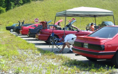 2007 Ford Mustang - Several Ford Mustangs are parked in a row at an outdoor meet, with one red convertible's trunk open.