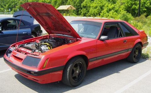 2007 Ford Mustang - Red 1985 Ford Mustang GT parked with its hood open, revealing the engine bay. Another vehicle is visible in the background.