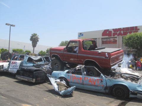 1978 Ford Bronco - A red 1978 Ford Bronco is perched atop a pile of wrecked cars, with a "4 Wheel Parts" store visible in the background.