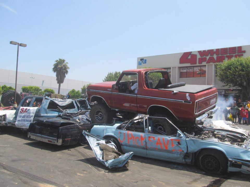 Red 1978 Ford Bronco on top of crushed cars, with "4 Wheel Parts" store in background