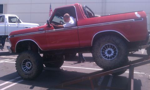 1978 Ford Bronco - Red 1978 Ford Bronco lifted on ramps with a driver visible. Another Ford truck is parked behind it.