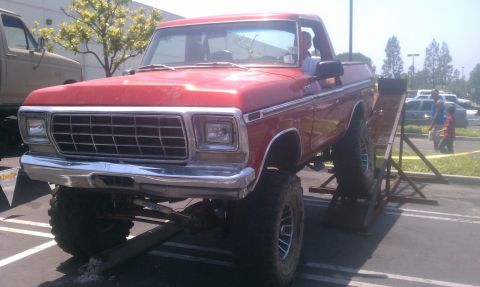 1978 Ford Bronco - A red pickup truck is positioned on ramps, with another vehicle visible to its left in a parking lot.