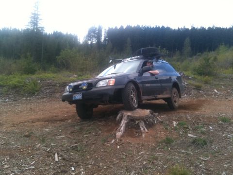 2005 Jeep Wrangler - A dark Subaru Outback station wagon navigates an off-road obstacle on a dirt path surrounded by trees.