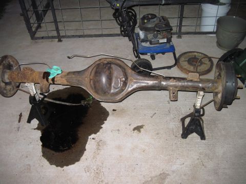 1979 Ford Bronco - A rusty rear axle assembly is supported by jack stands, with a dark oil spill on the concrete floor.